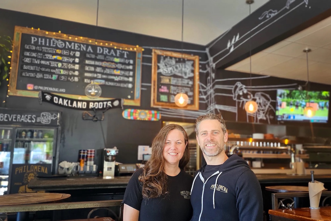 This photo shows the husband-and-wife owners of Philomena smiling at the camera with their restaurant's beer board in the background.