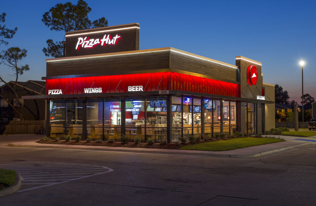 This photo shows the brightly lit exterior of a Pizza Hut restaurant against a dark blue sky.