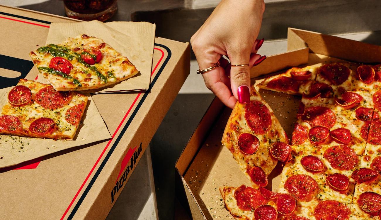 This photo shows a woman's hand removing a slice of tavern-style pizza from a whole pie in a Pizza Hut branded box.
