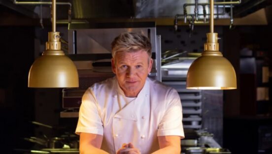 This photo shows Gordon Ramsay in a white chef coat, leaning forward against a table between two lamps hanging from the ceiling.