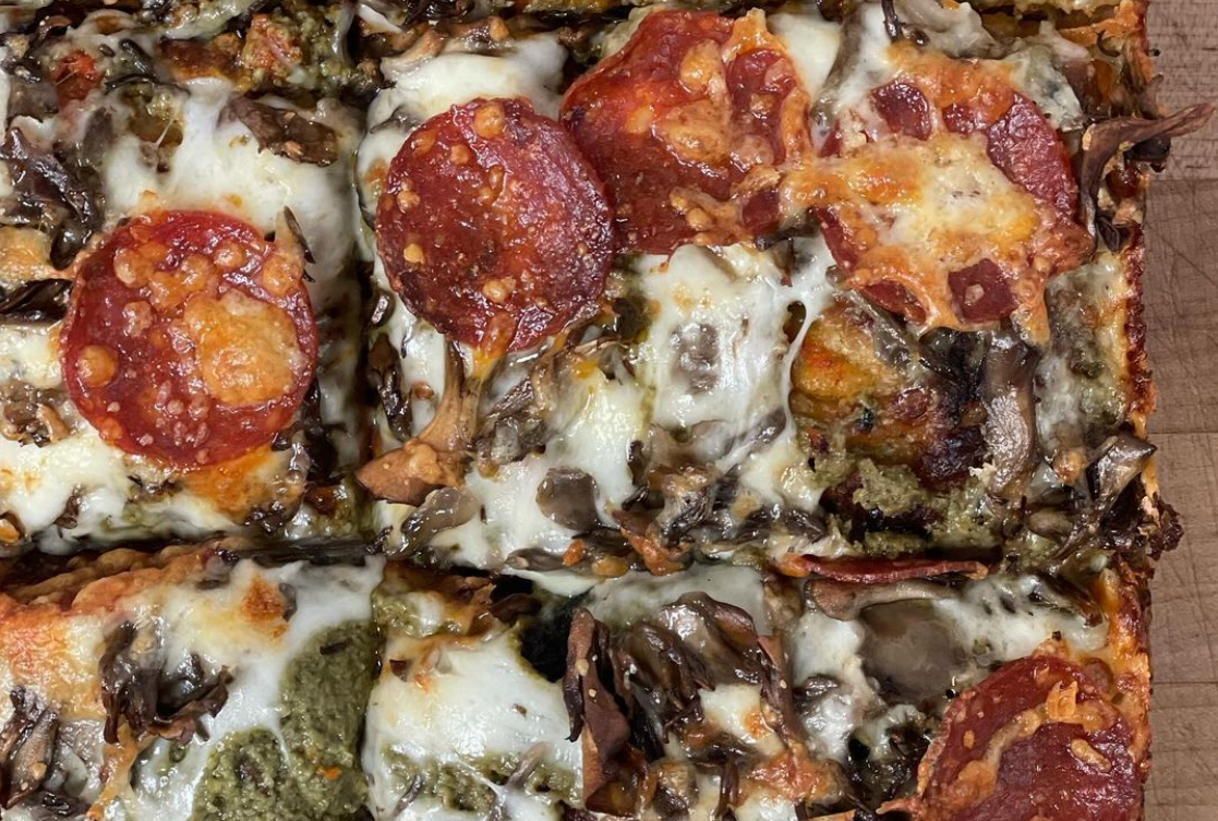This photo shows a close-up of a pizza that's a mix of Detroit and Sicilian styles, with pepperoni, mushrooms, mozzarella and what looks like perhaps a pesto.