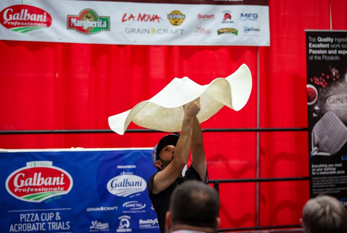 This photo shows a man spinning a large disk of dough in the air at a pizza competition.