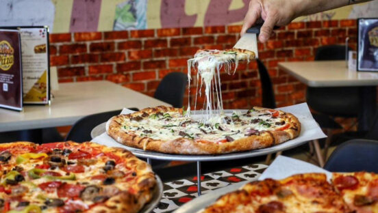 In this photo, a man with a tattooed arm is shown using a spatula to remove a slice of pizza with a great cheese melt from a whole pie, with two other pizzas nearby.