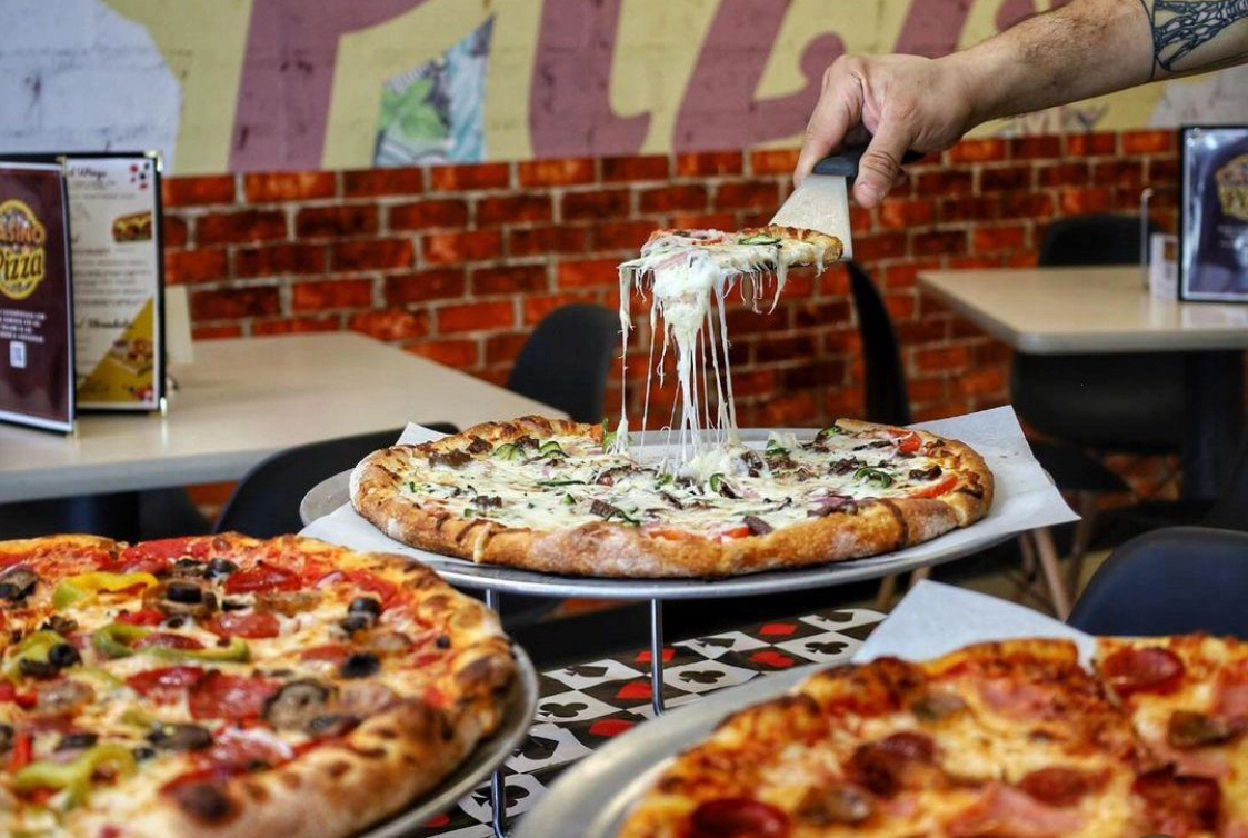 In this photo, a man with a tattooed arm is shown using a spatula to remove a slice of pizza with a great cheese melt from a whole pie, with two other pizzas nearby.