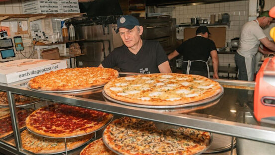 This photo shows Frank Kabatas, wearing a black shirt and cap, standing at the slice counter at East Village Pizza.