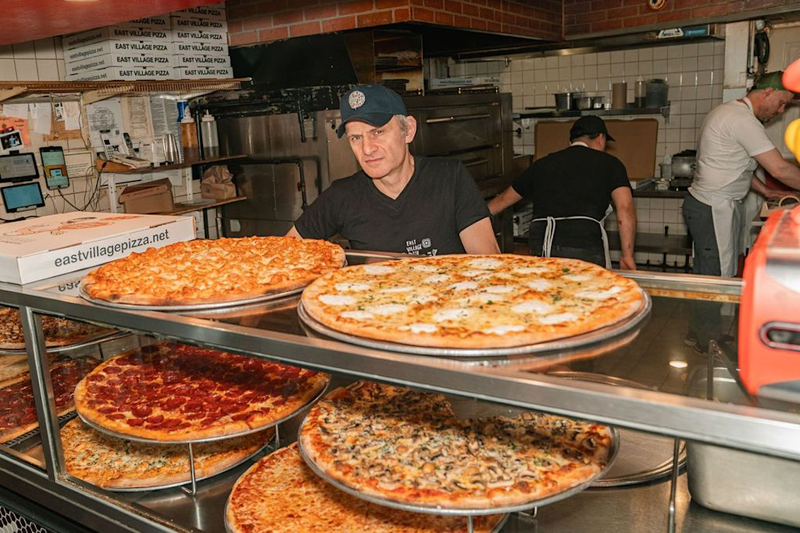 This photo shows Frank Kabatas, wearing a black shirt and cap, standing at the slice counter at East Village Pizza.