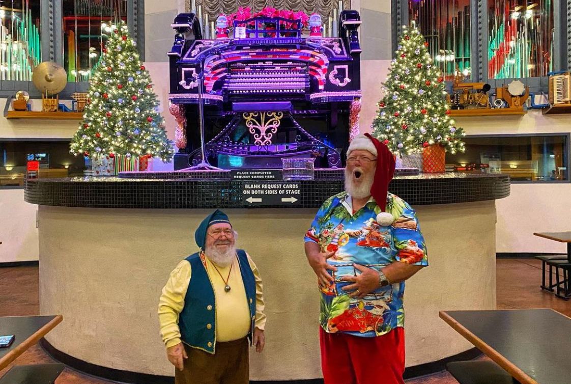 This photo shows a man in a Santa Claus hat and Hawaiian shirt, holding his belly and laughing like Santa Claus, along with a shorter man dressed as an elf.