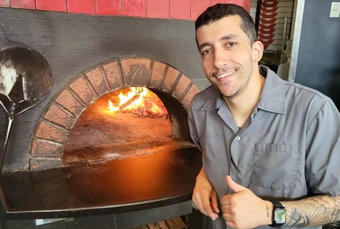 This photo shows Juan G Perez, with dark hair and wearing a gray shirt, smiling and giving the thumbs-up next to a wood-fired oven.