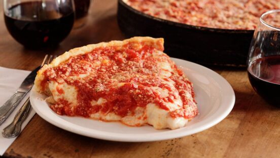 This photo shows a slice of Lou Malnati's deep-dish pizza on a plate with a whole pizza in a pan off to the right.