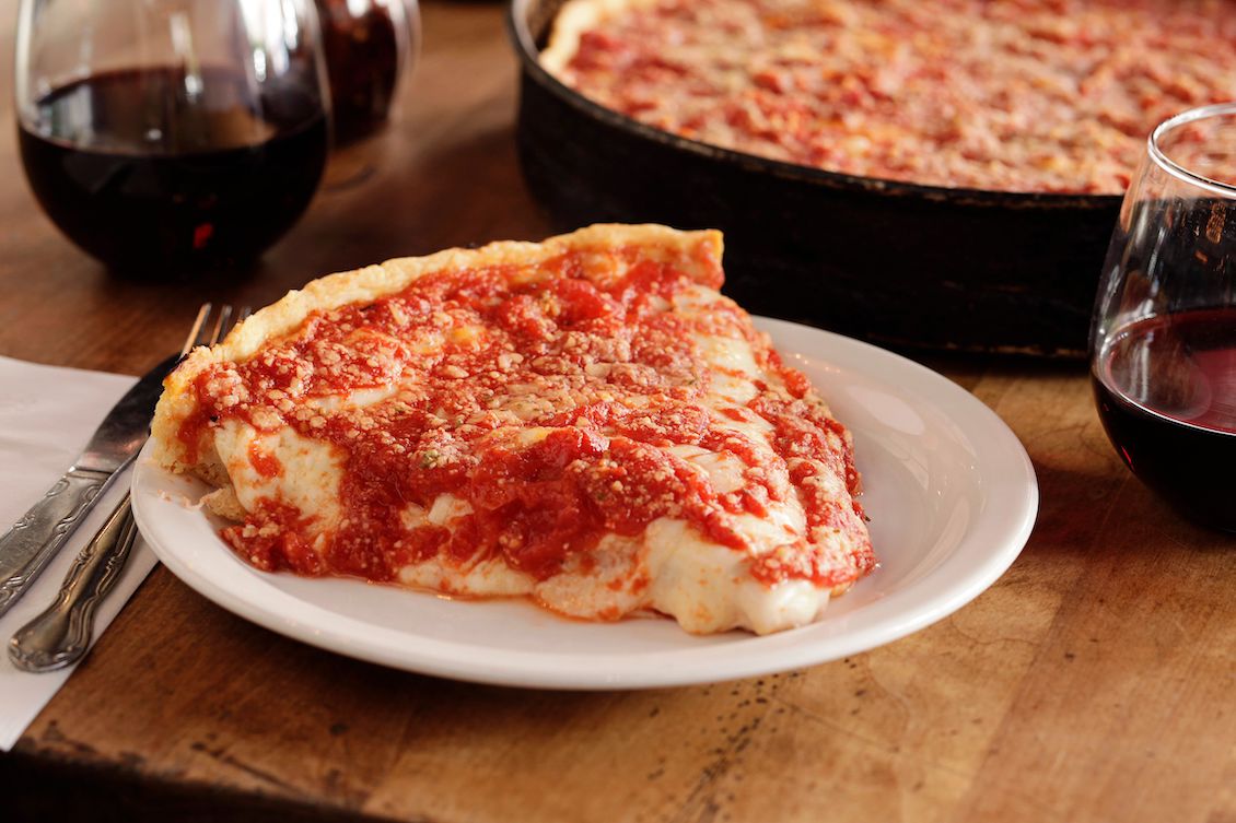 This photo shows a slice of Lou Malnati's deep-dish pizza on a plate with a whole pizza in a pan off to the right.