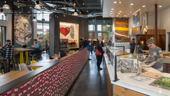 This photo shows the interior of a MOD Pizza restaurant, with customers waiting for pizza and a couple of employees making pies.
