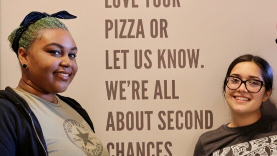 This photo shows two female MOD Pizza employees standing in front of a wall that has writing on it: Love your pizza or let us know. We're all about second chances."