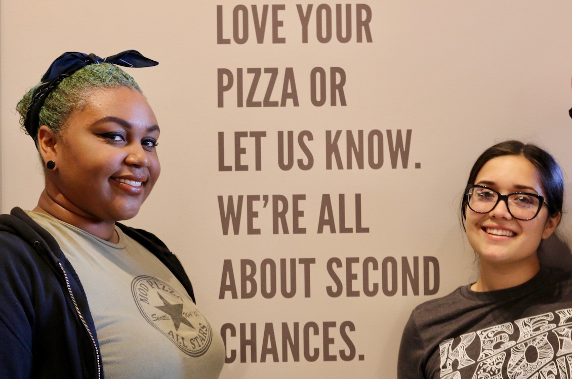 This photo shows two female MOD Pizza employees standing in front of a wall that has writing on it: Love your pizza or let us know. We're all about second chances."
