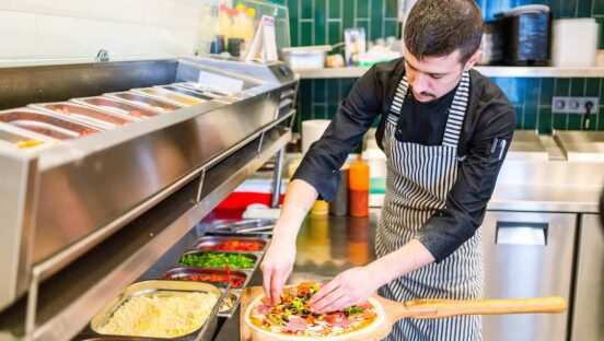 This photo shows a man with dark hair and a mustache and goatee making pizza in a busy restaurant kitchen.