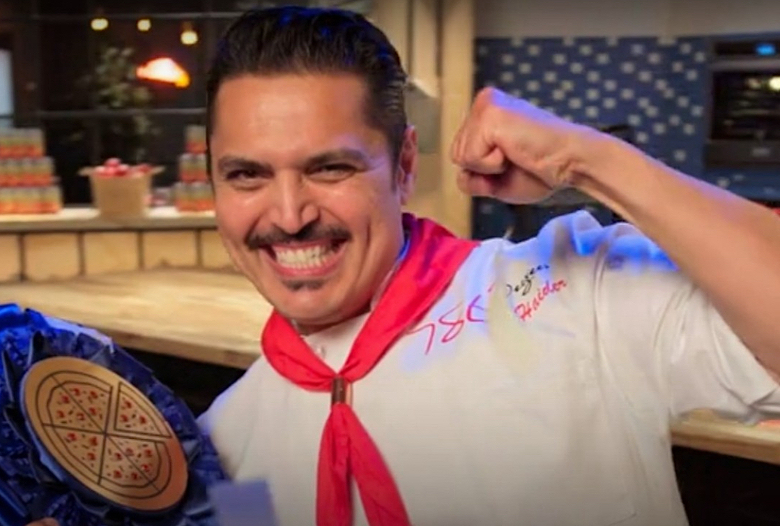 This photo shows U.S. Pizza Team member Ali Haider, wearing a white chef coat with a red bandana, smiling hugely and making a fist.