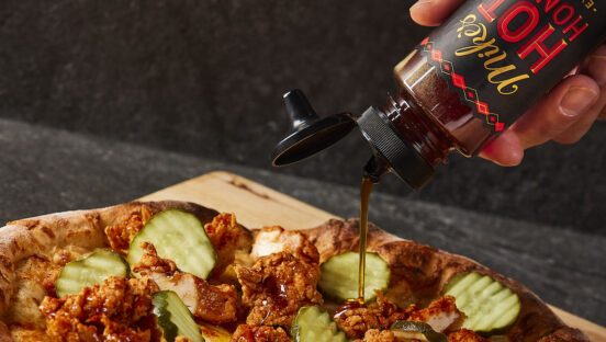 This photo shows Mike's Hot Honey being drizzled over a pizza topped with Nashville hot chicken, mozzarella, dill pickle slices and other ingredients.