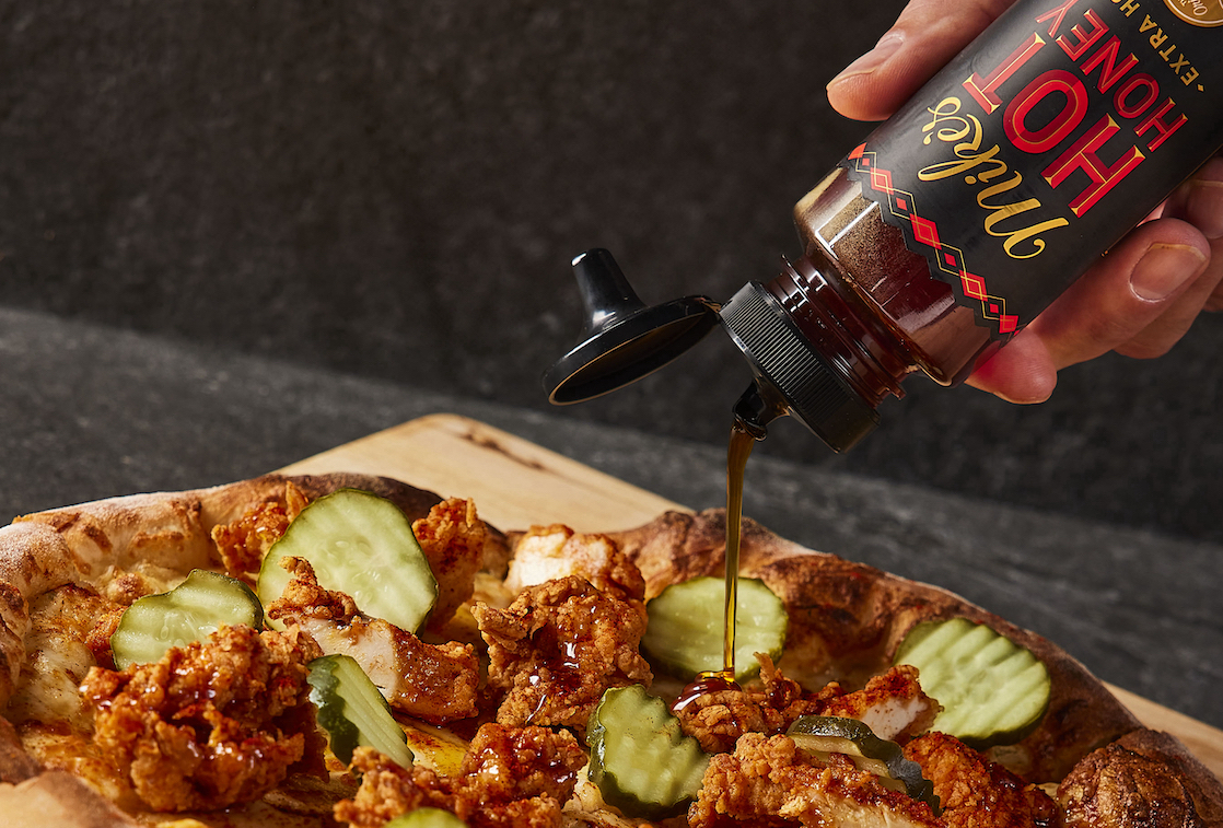 This photo shows Mike's Hot Honey being drizzled over a pizza topped with Nashville hot chicken, mozzarella, dill pickle slices and other ingredients.