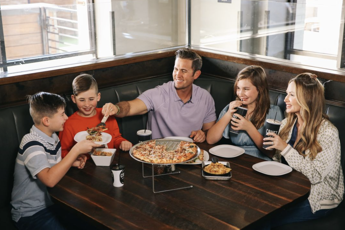 A happy family of five digs into a shared pizza at a corner booth.