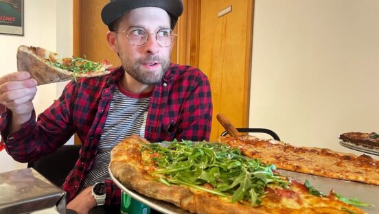 This photo shows Dan Tallarico, wearing a black cap and a checkered shirt, getting ready to eat a slice of pizza, with a couple of large whole pies in front of him.