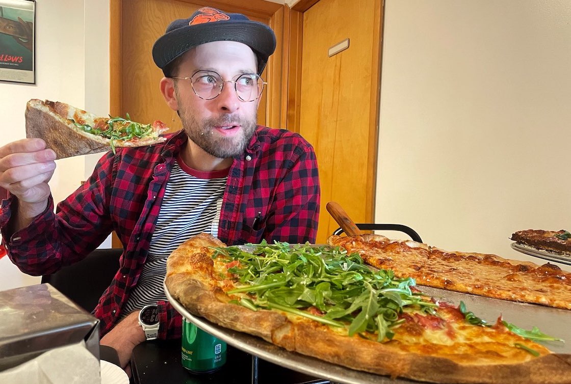 This photo shows Dan Tallarico, wearing a black cap and a checkered shirt, getting ready to eat a slice of pizza, with a couple of large whole pies in front of him.