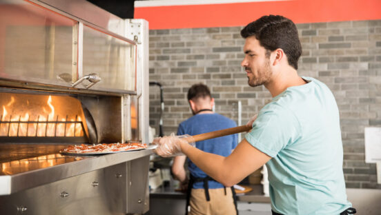 Side view of a young chef using peel to put pizza in oven for baking