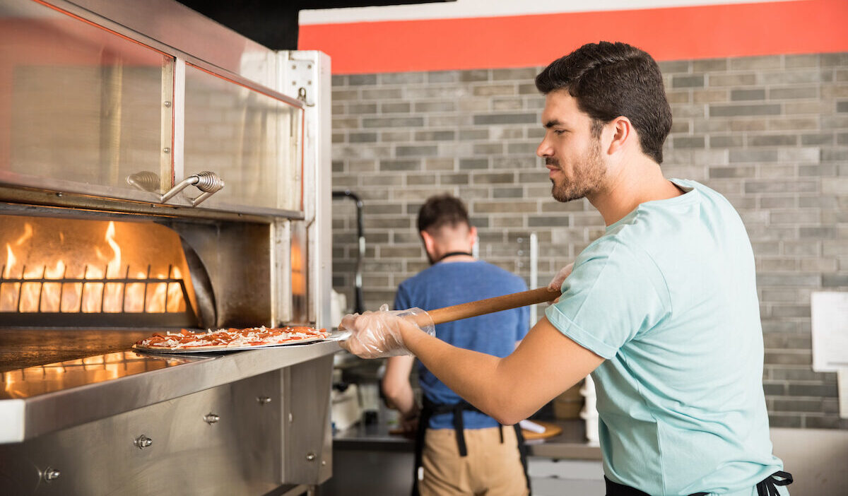 Side view of a young chef using peel to put pizza in oven for baking