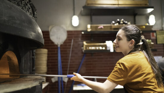 This photo shows a pony-tailed woman peeling a pizza into an oven in a restaurant kitchen.