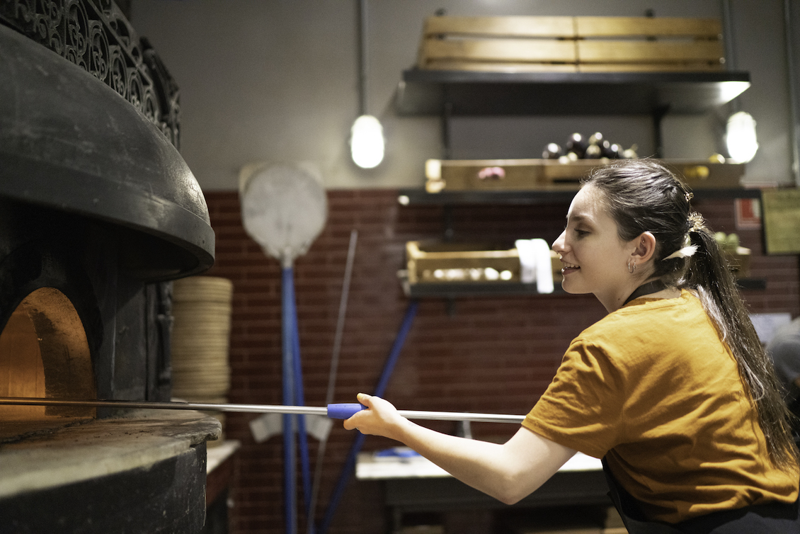 This photo shows a pony-tailed woman peeling a pizza into an oven in a restaurant kitchen.