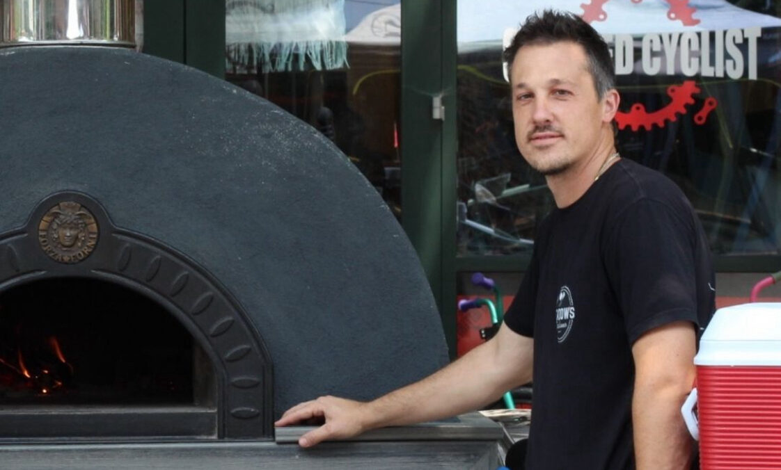 This photo shows the late Jayson Poluka, wearing a dark shirt, standing in front of a wood-fired pizza oven.