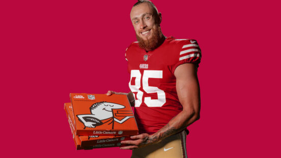 This photo shows San Francisco 49ers George Kittle in his football uniform, posing against a dark red background with a box of Little Caesars pizza in his hands.