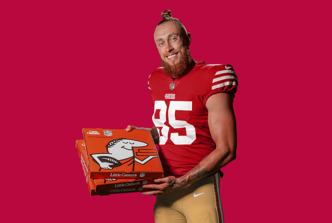This photo shows San Francisco 49ers George Kittle in his football uniform, posing against a dark red background with a box of Little Caesars pizza in his hands.