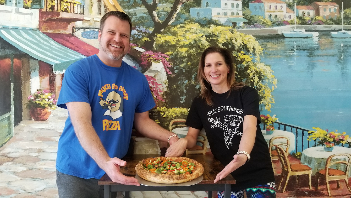 This photo shows Mark and Kira proudly presenting one of their pizzas, which is laid on a table between them.