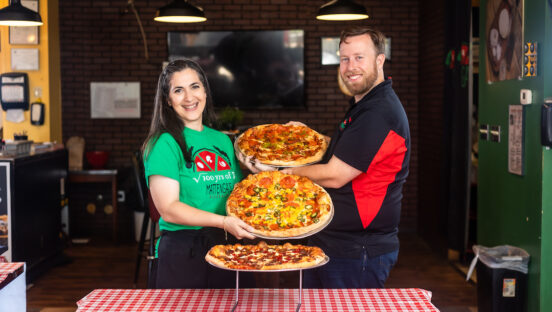 This photo shows Hengam Stanfield (wearing a bright green t-shirt) and Matt Stanfield (in a dark blue and red shirt) standing in front of a checkered-cloth table. Between them are three pizzas with various toppings.