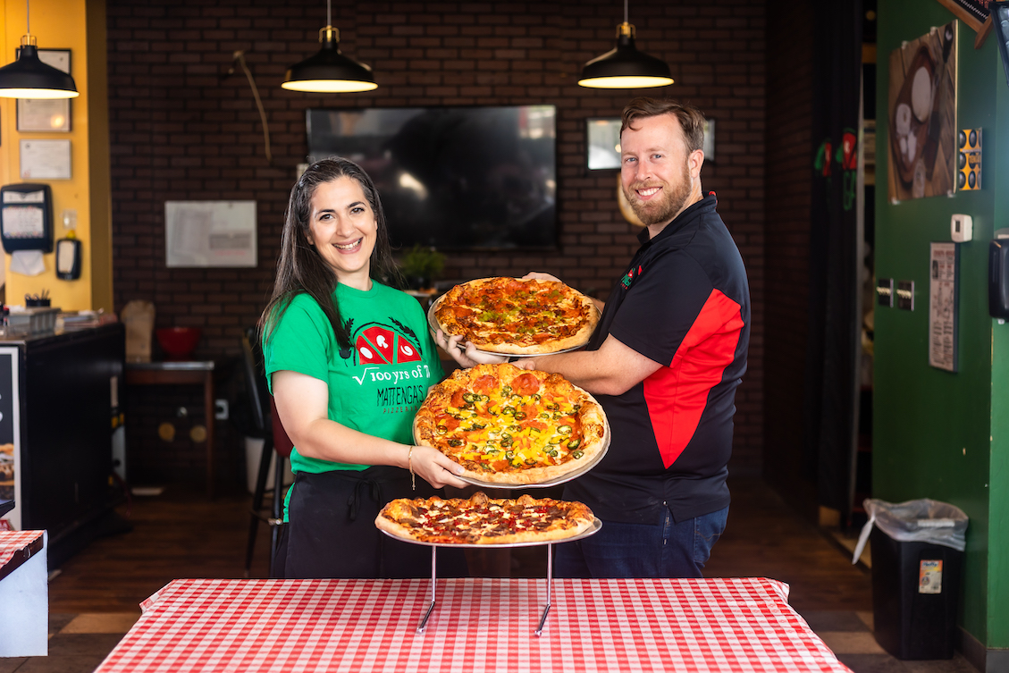 This photo shows Hengam Stanfield (wearing a bright green t-shirt) and Matt Stanfield (in a dark blue and red shirt) standing in front of a checkered-cloth table. Between them are three pizzas with various toppings.