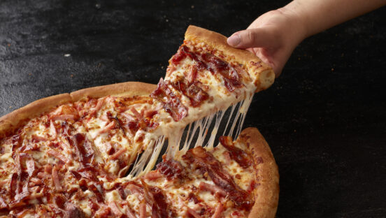 This photo shows a person's hand pulling out a slice from a whole Triple Bacon pie with a solid cheese pull.