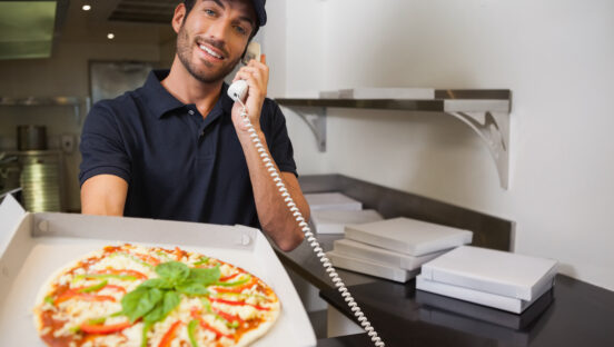This photo shows a lightly bearded man in a dark shirt and cap, holding out a pizza and talking on the phone.