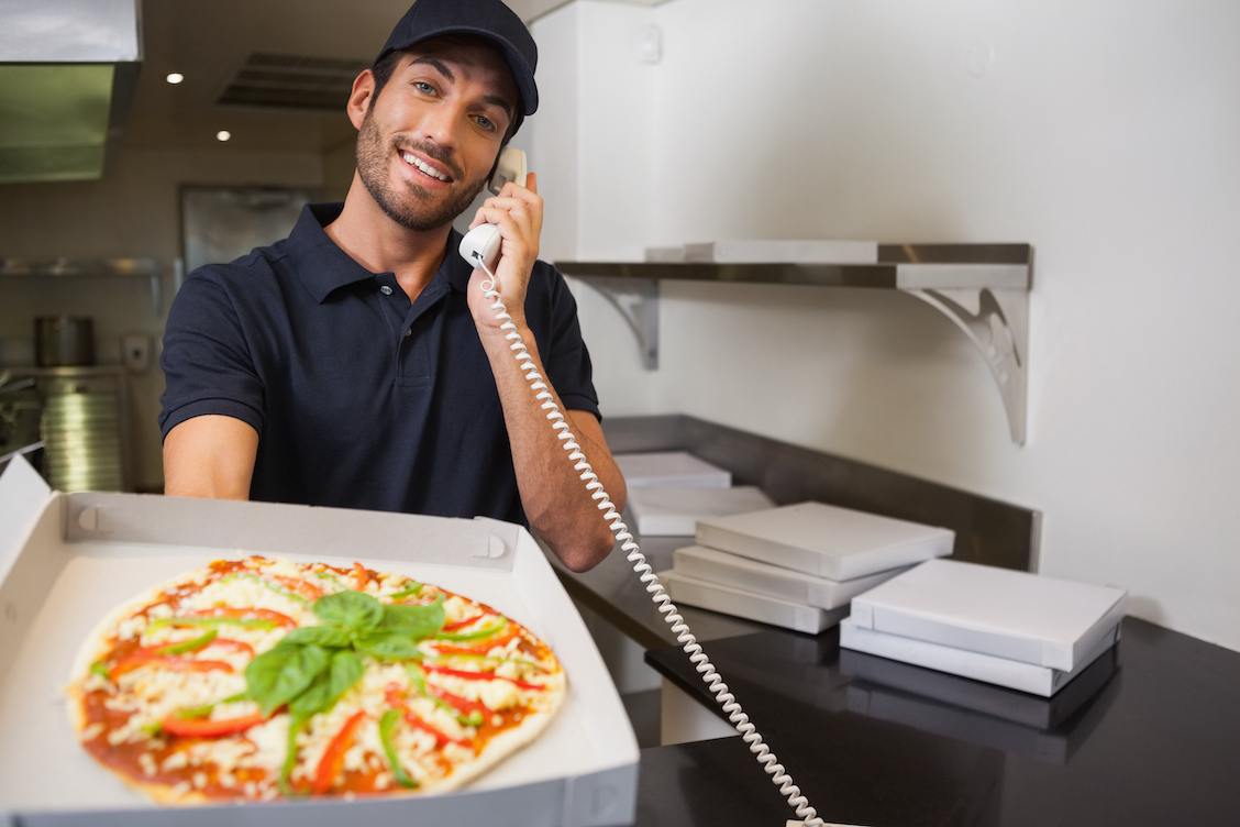 This photo shows a lightly bearded man in a dark shirt and cap, holding out a pizza and talking on the phone.