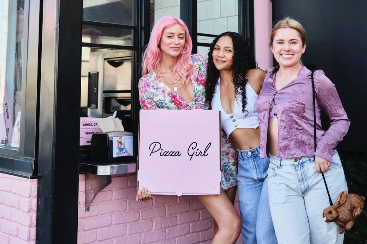 three women stand in front of a pizzeria, with pink brick walls, holding a pizza box that says "Pizza Girl."
