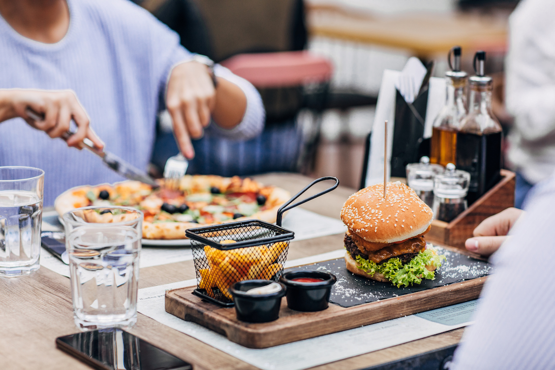 This photo shows two people eating at a restaurant. One is cutting into a pizza with a knife and fork while a hamburger and fries sit on a tray in front of the other person.