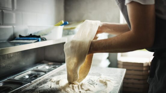 This photo shows a Closeup side view of a chef stretching pizza dough at a local pizza place.