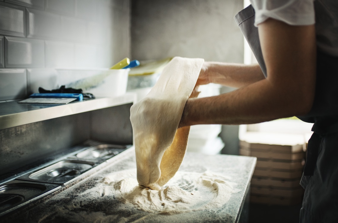 This photo shows a Closeup side view of a chef stretching pizza dough at a local pizza place.