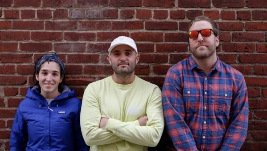 This photo shows the three owners - a woman and two men - of Timber Pizza Co. standing side by side against a red-brick wall.