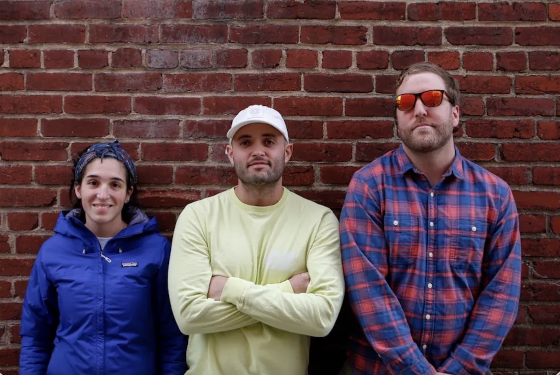 This photo shows the three owners - a woman and two men - of Timber Pizza Co. standing side by side against a red-brick wall.