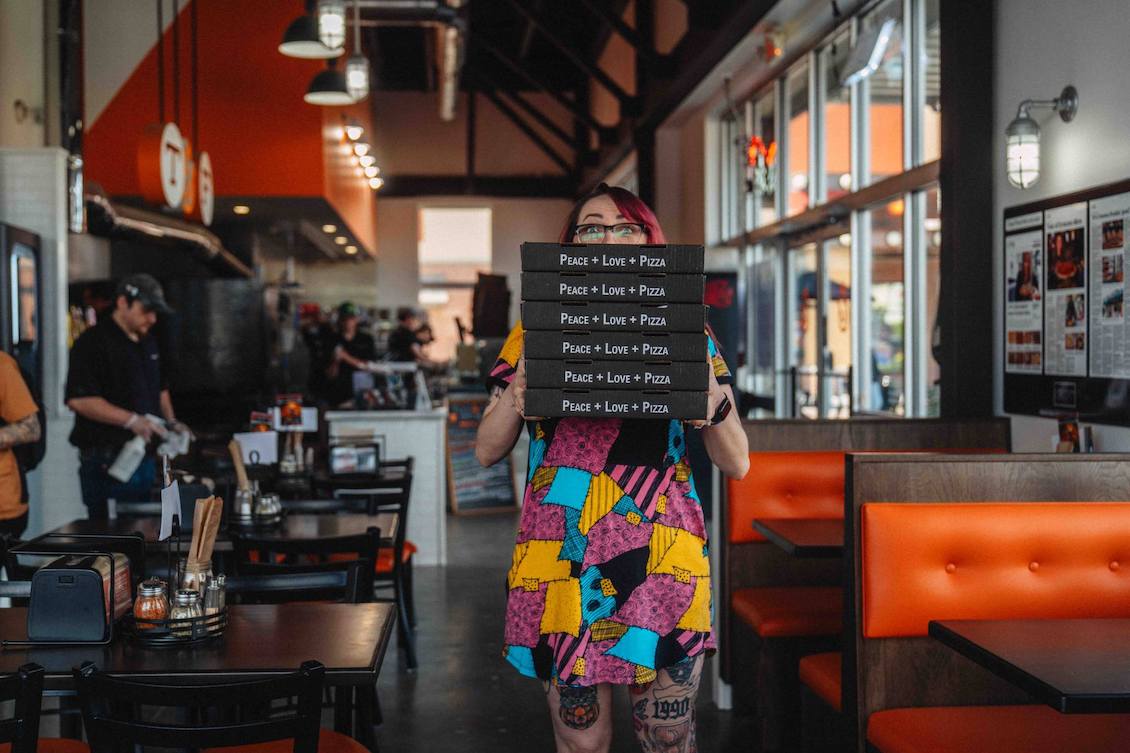 This photo shows a woman standing in the dining room of a Toss & Fire restaurant, holding a stack of black pizza boxes with white lettering that reads "Peace + Love + Pizza."