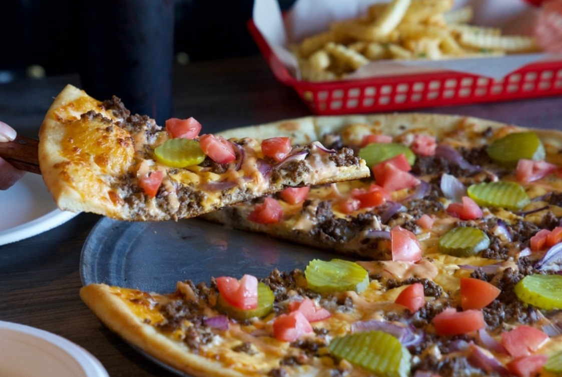 This photo shows a cheeseburger pizza at Village Pizzeria, topped with ground beef, tomatoes, pickles and cheddar and mozzarella cheeses.