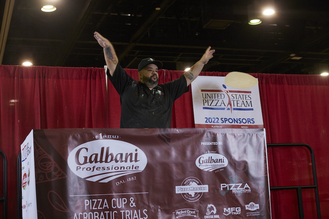 This photo shows Wilhelm Rodriguez, in a black U.S. Pizza Team chef coat, raising his hands triumphantly at a pizza competition.