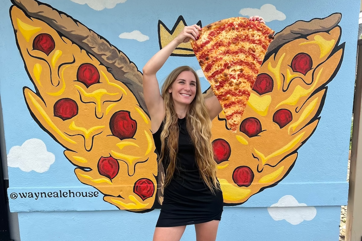 A woman stands in front of a mural with pizza-themed butterfly wings holding up a piece of pizza.