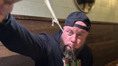 A bearded man with a backwards hat and a shirt that says The Pizza Hulk holds a slice of pizza high as a string of cheese leaves his mouth.