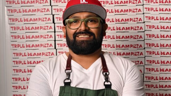 A man stands in an apron donning a red Dodgers hat. Logos behind him say "Triple Beam Pizza."