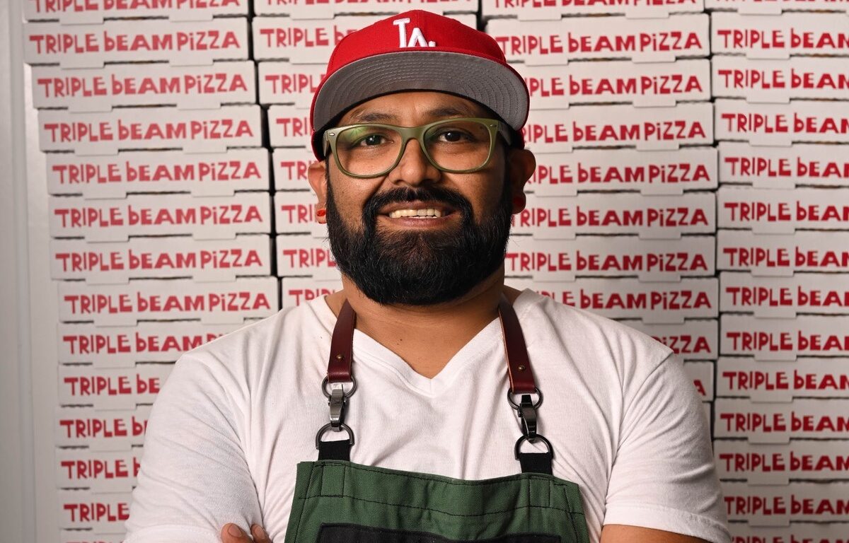 A man stands in an apron donning a red Dodgers hat. Logos behind him say "Triple Beam Pizza."